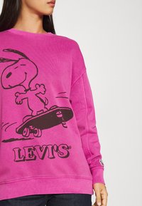 Sudadera rosa con un gráfico negro de Snoopy patinando. Logo "LEVI'S" en negro en la parte inferior. Textura suave con un ajuste holgado.