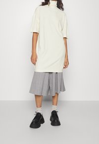 Lys beige oversized turtleneck kjole med korte ærmer, parret med en grå stribet plisseret nederdel og sorte platformsko.