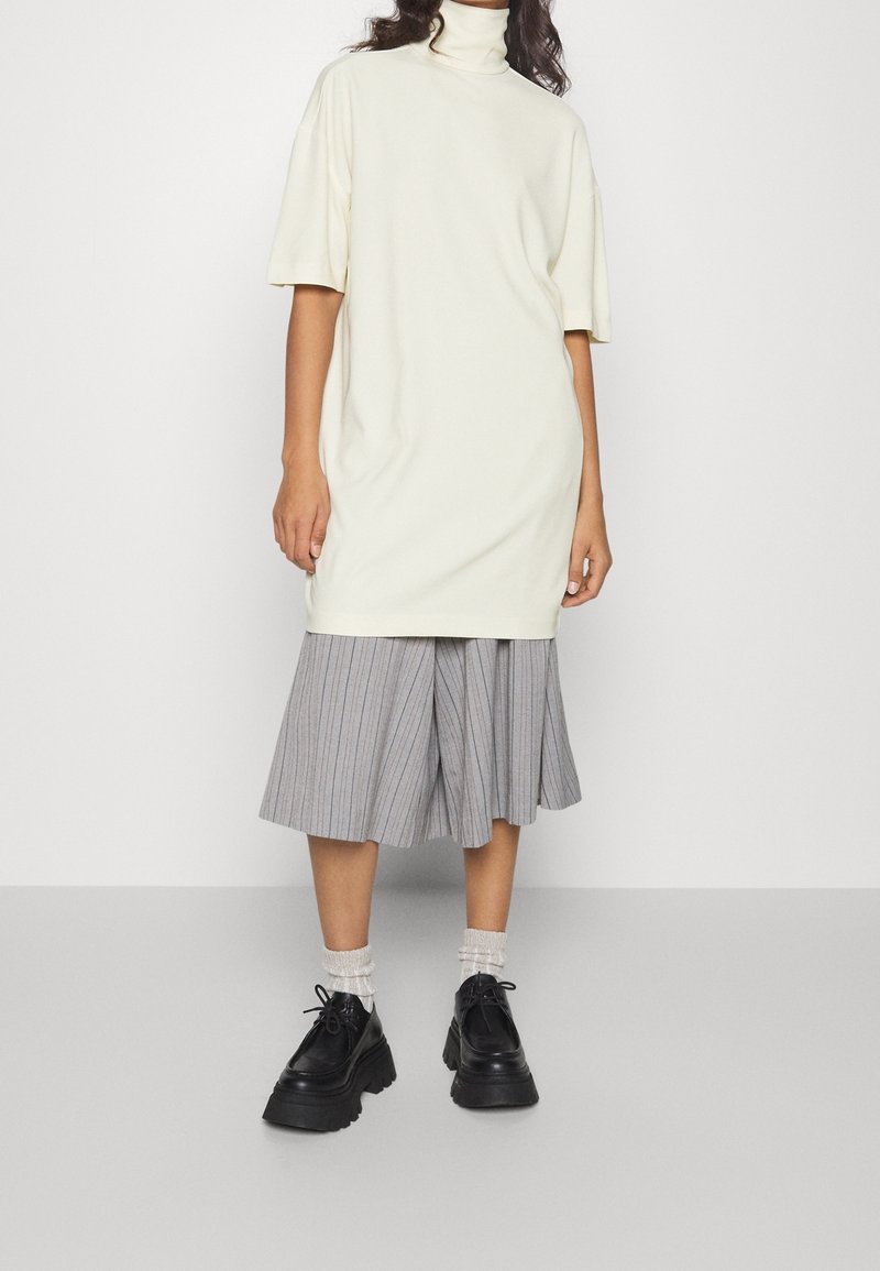 Lys beige oversized turtleneck kjole med korte ærmer, parret med en grå stribet plisseret nederdel og sorte platformsko.