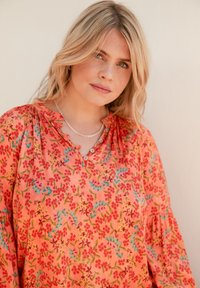 Blusa floreale in corallo con fiori rossi vivaci e foglie verdi. Presenta un colletto con bottoni e maniche a sbuffo, realizzata in tessuto leggero.