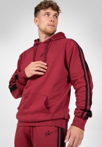 Bordeauxkleurige hoodie met zwarte zijstrepen, een capuchon met trekkoord, een buidelzak en logodetail. Zachte textuur, ontspannen pasvorm.