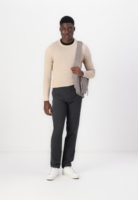 Beige gewatteerde trui, grijze broek en witte sneakers. Grijze schoudertas met ritsdetail. Effen witte achtergrond.