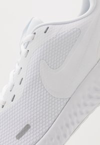 Zapato deportivo blanco con una parte superior de malla texturizada, acentos lisos y una suela de goma con un patrón ondulado. Diseño de cordones y detalle del logo.