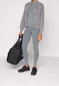 Giacca bomber grigia con polsini a costine, top grigio aderente abbinato e leggings; scarpe da ginnastica nere; tiene una borsa a tracolla nera.