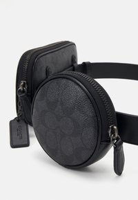 Svart dubbelkompartmentsväska med en texturerad rund bas och en slät rektangulär topp, båda med blixtlås och en avtagbar tag.