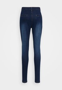 Jeans skinny de tiro alto en un azul oscuro, con dos bolsillos traseros con ribete y costuras en contraste, mostrados sobre un fondo gris claro.