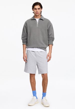 Jeune homme debout portant un pull gris à fermeture éclair quart de zip, un t-shirt blanc, un short gris clair, des chaussettes bleues et des baskets blanches sur fond blanc.