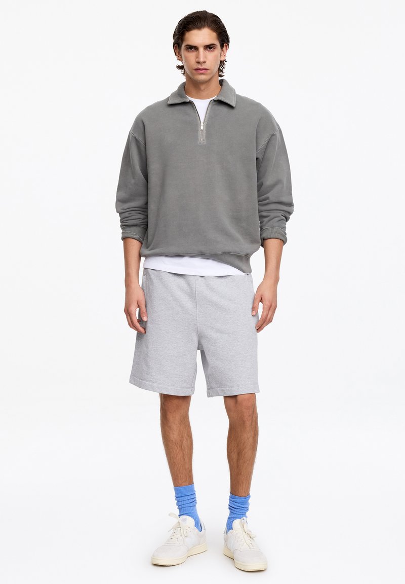 Jeune homme debout portant un pull gris à fermeture éclair quart de zip, un t-shirt blanc, un short gris clair, des chaussettes bleues et des baskets blanches sur fond blanc.