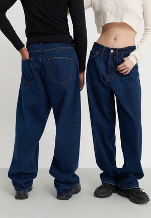 Par löst sittande mörkblå jeans i denim med fram- och bakfickor, med hög midja och uppvikta fållar. Bärs med svarta skor.