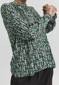Blusa floreale verde, nera e bianca con dettagli viola. Presenta un collo alto, un design plissettato e maniche lunghe con polsini arricciati.