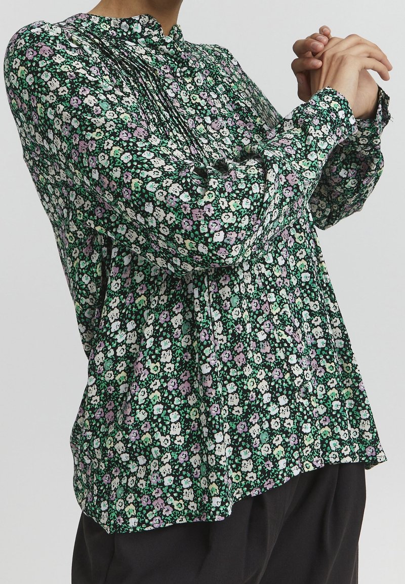 Blusa floreale verde, nera e bianca con dettagli viola. Presenta un collo alto, un design plissettato e maniche lunghe con polsini arricciati.