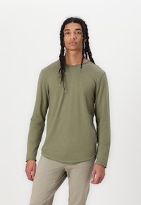 Langarm-Hemd in Olivgrün, mit abgerundetem Saum, weichem Textil und rohen Kantendetails. Kombiniert mit hellgrauen, taillierten Hosen.
