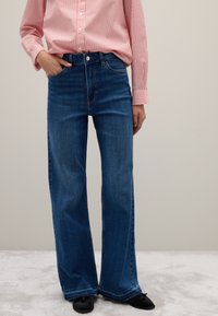 Donna che indossa jeans blu a vita alta e gamba larga, una camicia a righe rosa e bianche con una mano in tasca, in piedi su un tappeto grigio chiaro.
