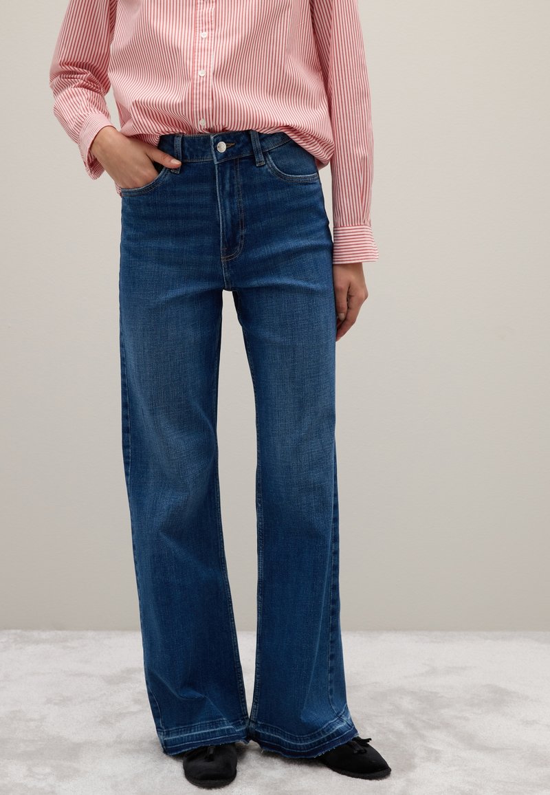 Donna che indossa jeans blu a vita alta e gamba larga, una camicia a righe rosa e bianche con una mano in tasca, in piedi su un tappeto grigio chiaro.