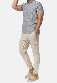 Kurzärmliges Hemd mit Knöpfen, grau-weiß gestreift, kombiniert mit beigen Hosen mit elastischen Bündchen und hellen Schlappen.