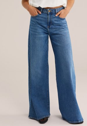 Femme portant un jean bleu taille haute à jambes larges, les mains dans les poches et un haut blanc visible à la hauteur de la taille.