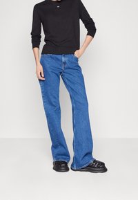 Vaqueros de mezclilla azules acampanados, de cintura alta y ajuste regular. Combinados con una blusa negra de manga larga y unos zapatos negros de plataforma sin cordones.