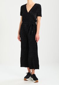 Svart jumpsuit med korta ärmar, diskreta vertikala ränder, knytning i midjan och vida ben, kombinerad med svarta sportskor.