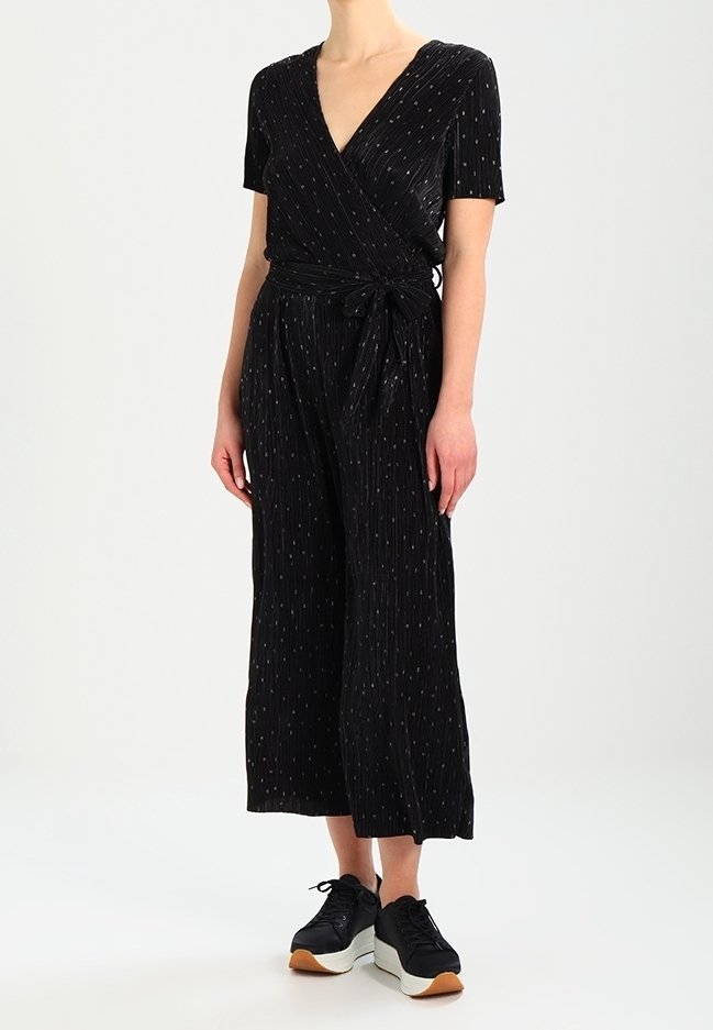 Svart jumpsuit med korta ärmar, diskreta vertikala ränder, knytning i midjan och vida ben, kombinerad med svarta sportskor.