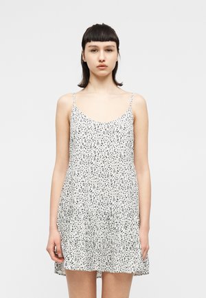 Tânără cu păr drept și închis la culoare, purtând o rochie mini albă, fără mâneci, cu imprimeu floral negru, stând pe fundal gri deschis.