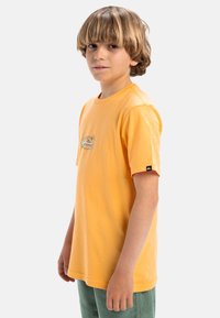 Camiseta de algodón amarilla de manga corta, con un pequeño logotipo gráfico y una etiqueta en la manga izquierda. Combinada con pantalones de pana verde.