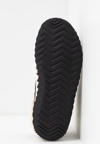 Semelle de chaussure en caoutchouc noire avec un motif de bande de roulement en chevrons profonds, positionnée à la verticale contre un coin blanc en arrière-plan.