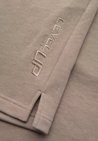 Tissu beige avec texte brodé « LEVEL UP » et une petite fente latérale sur l’ourlet d’un vêtement.