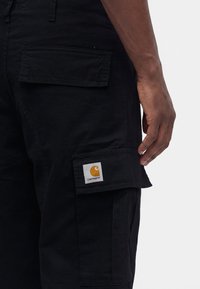 Gros plan sur un pantalon cargo noir avec des poches à rabats et un patch logo Carhartt jaune sur la poche inférieure, porté par une personne à la peau foncée.