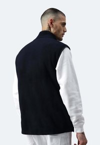 Marinefarbener Fleece-Weste ohne Ärmel mit hohem Kragen. Getragen über einem weißen Langarmhemd, mit sichtbaren Tätowierungen am Arm.