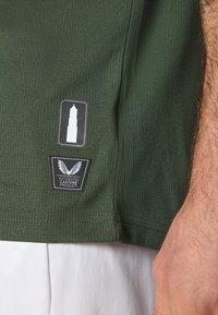 Olīvu zaļš teksturēts T-krekls ar birku, kurai ir balts torņa grafiks un melns uzraksts "AUTENTISKS CASTORE PRODUKTS" apakšmalā.