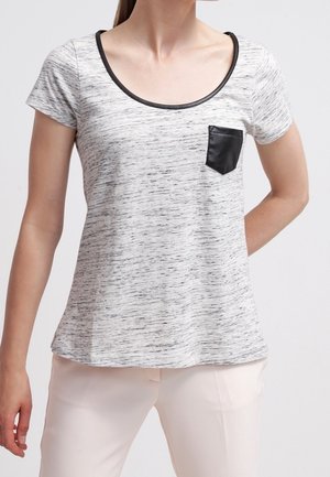 Woman wearing a short-sleeve light gray heathered top with black trim neckline and black faux leather chest pocket, paired with light pants.