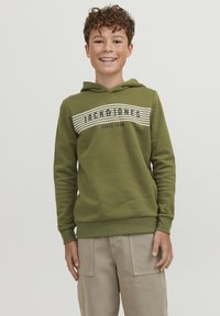 Sudadera verde con capucha y un diseño de franjas blancas en el frontal con texto. Puños y dobladillo acanalados, hecha de un tejido suave. Llevada con pantalones cargo de color beige claro.