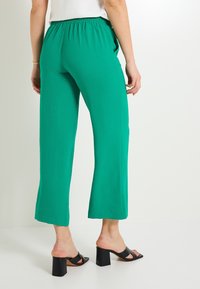 Pantalones verdes de pierna ancha con cinturilla elástica, hechos de una tela texturizada. Combinados con sandalias negras de tacón bloque.