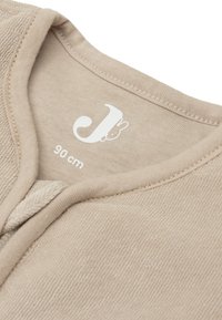 Tissu de vêtement beige clair avec une surface texturée, présentant un col rond et une étiquette blanche imprimée indiquant la taille (90 cm).