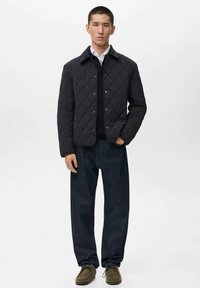 Chaqueta oscura acolchada con botones a presión, usada sobre un suéter negro y una camisa blanca, combinada con pantalones vaqueros oscuros holgados y zapatos marrones.