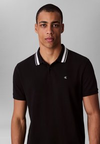 Black polo shirt featuring a white-striped collar, logo on the chest, short sleeves, and smooth cotton fabric. Plain background.