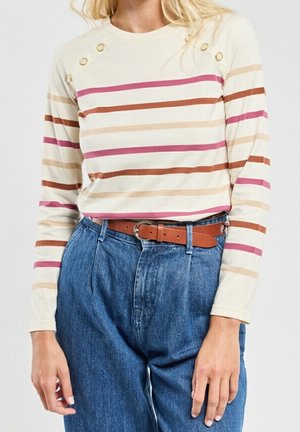 Kobieta w kremowej bluzce z długim rękawem, ozdobionej wielokolorowymi poziomymi paskami i złotymi guzikami na ramieniu, wpuszczonej w wysokiej talii niebieskie dżinsy z brązowym paskiem.