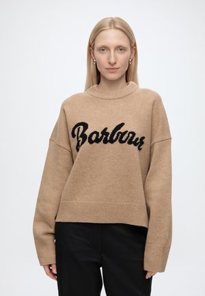 Blondynka ubrana w beżowy sweter z dzianiny z czarnym napisem "Barbour", w połączeniu z czarnymi spodniami, stojąca na jednolitym jasnym tle.