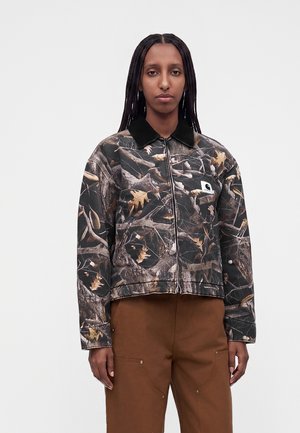 Femme aux longues tresses portant une veste zippée à motif camouflage avec un col noir et un pantalon marron, debout devant un fond clair uni.