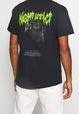 Person wearing black t-shirt with graphic of black bear and neon green "NIGHT ADDICT" text on back, paired with white pants.