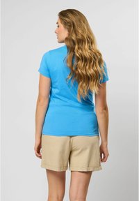 Blauwe katoenen T-shirt met korte mouwen, strakke pasvorm. Gecombineerd met beige shorts met opgerolde manchetten, tegen een neutrale achtergrond.