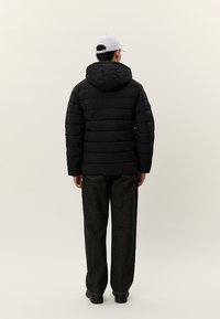 Mann iført en svart quiltet hettejakke, mørke løse jeans, svarte sko og en hvit baseballcaps, sett bakfra mot en ensfarget bakgrunn.