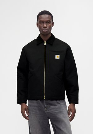 Homme portant une veste noire zippée avec un col et un petit logo Carhartt sur la poitrine ainsi qu’un jean gris sur un fond uni.