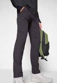 Sorte syntetiske bukser med slim fit, med lommer med glidelås og synlige sømmer. En grønn og svart ryggsekk holdes.