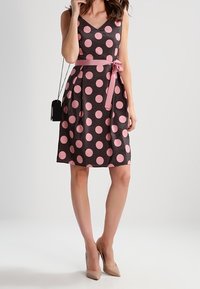 Vestido sin mangas de escote en V en marrón oscuro con grandes lunares rosas, cintura ajustada con un cinturón rosa, tejido suave, de longitud por encima de la rodilla.