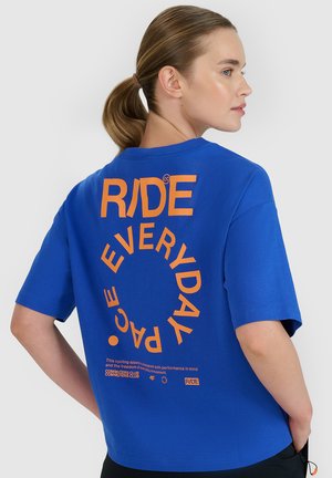 Kobieta z kucykiem ubrana w niebieską koszulkę z pomarańczowym napisem "RIDE EVERYDAY" ułożonym w okrągły wzór na plecach.