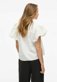 Blusa bianca a maniche corte con maniche arricciate, caratterizzata da una vestibilità rilassata, texture morbida e finitura liscia. Abbinata a pantaloni neri a gamba larga.