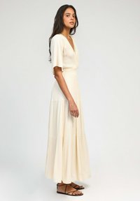 Robe maxi de couleur crème avec des manches courtes à volants, un décolleté en V et un design à volants, fabriquée en tissu texturé. Portée avec des sandales plates.