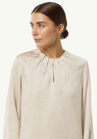 Hellbeige Bluse mit langen Ärmeln, rundem Ausschnitt und dezenten Falten am Halsauschnitt, aus glattem, strukturiertem Stoff.