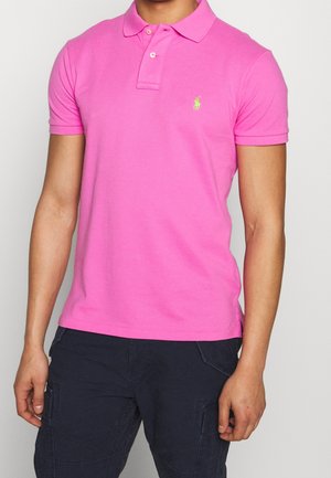 Mann trägt ein knallpinker kurzärmeliger Poloshirt mit dunkler Hose vor einem schlichten Hintergrund.